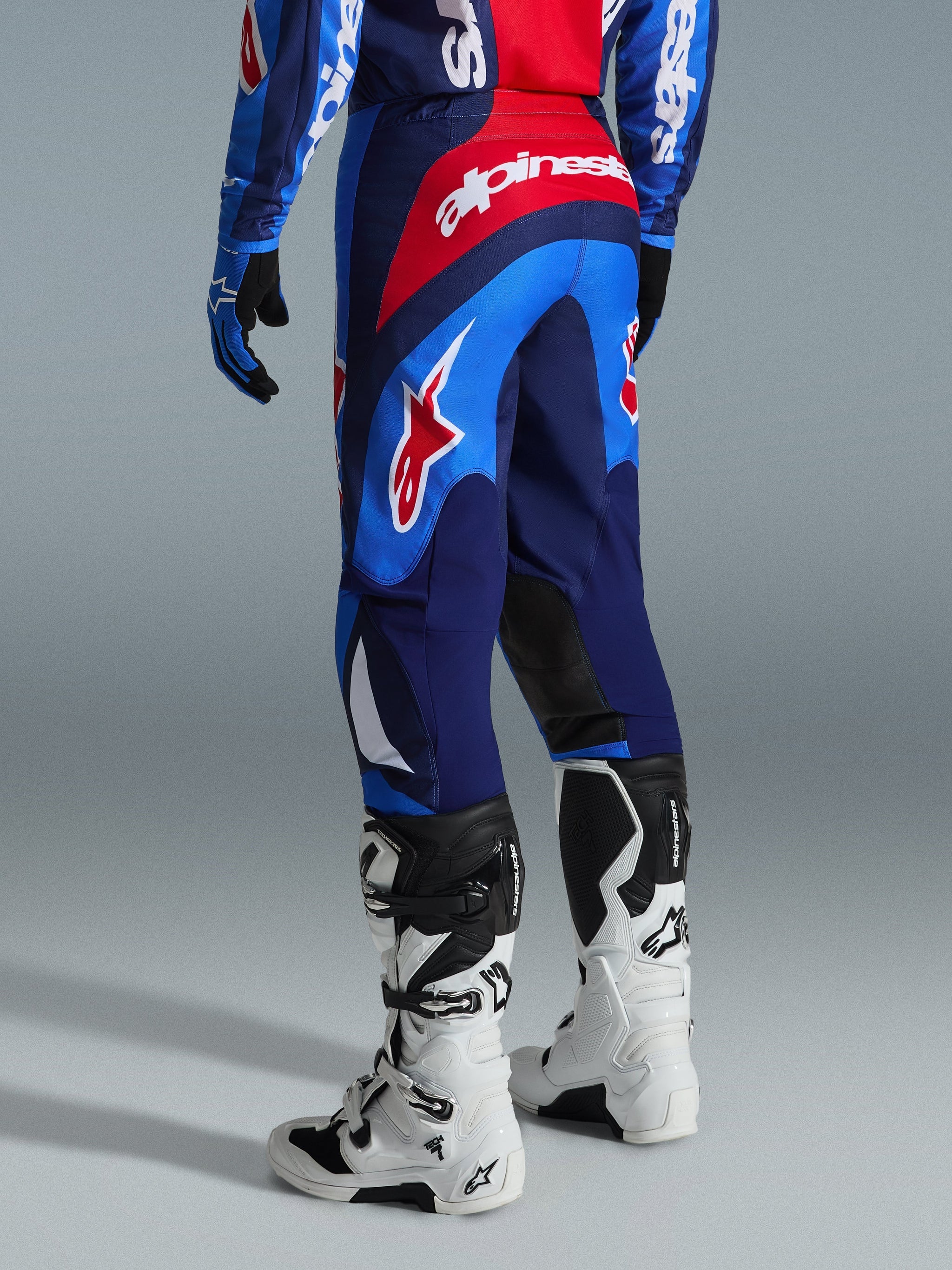 Alpinestars 2026 Fluid Grid Hose, Motocross-Hose, Blau und Rot, Rückansicht mit markantem Alpinestars-Branding und ergonomischem Design, getragen mit weiß-schwarzen Tech 7 Stiefeln und passendem Trikot, Hochleistungsausrüstung für das Offroad-Fahren