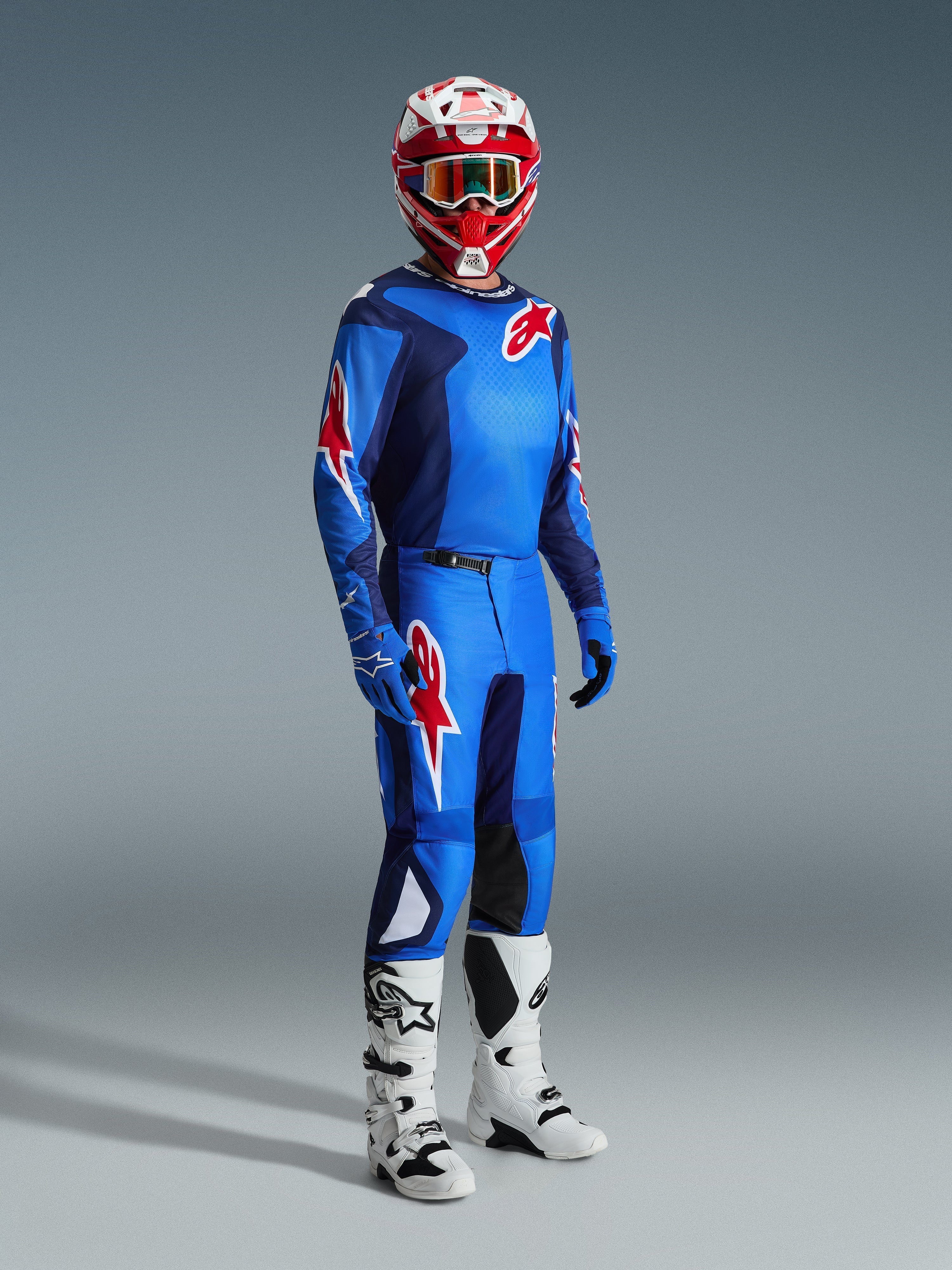 Motocross-Fahrer in kompletter Rennausrüstung, blau-rotes Trikot mit Gittermuster und Alpinestars-Logo, 2026 Fluid Grid Hose in Blau-Rot mit schwarzen Knieinnenseiten, rot-weißer Helm mit Schutzbrille, weiß-schwarze Stiefel, blaue Handschuhe, abgestimmtes Outfit für Offroad-Rennsport