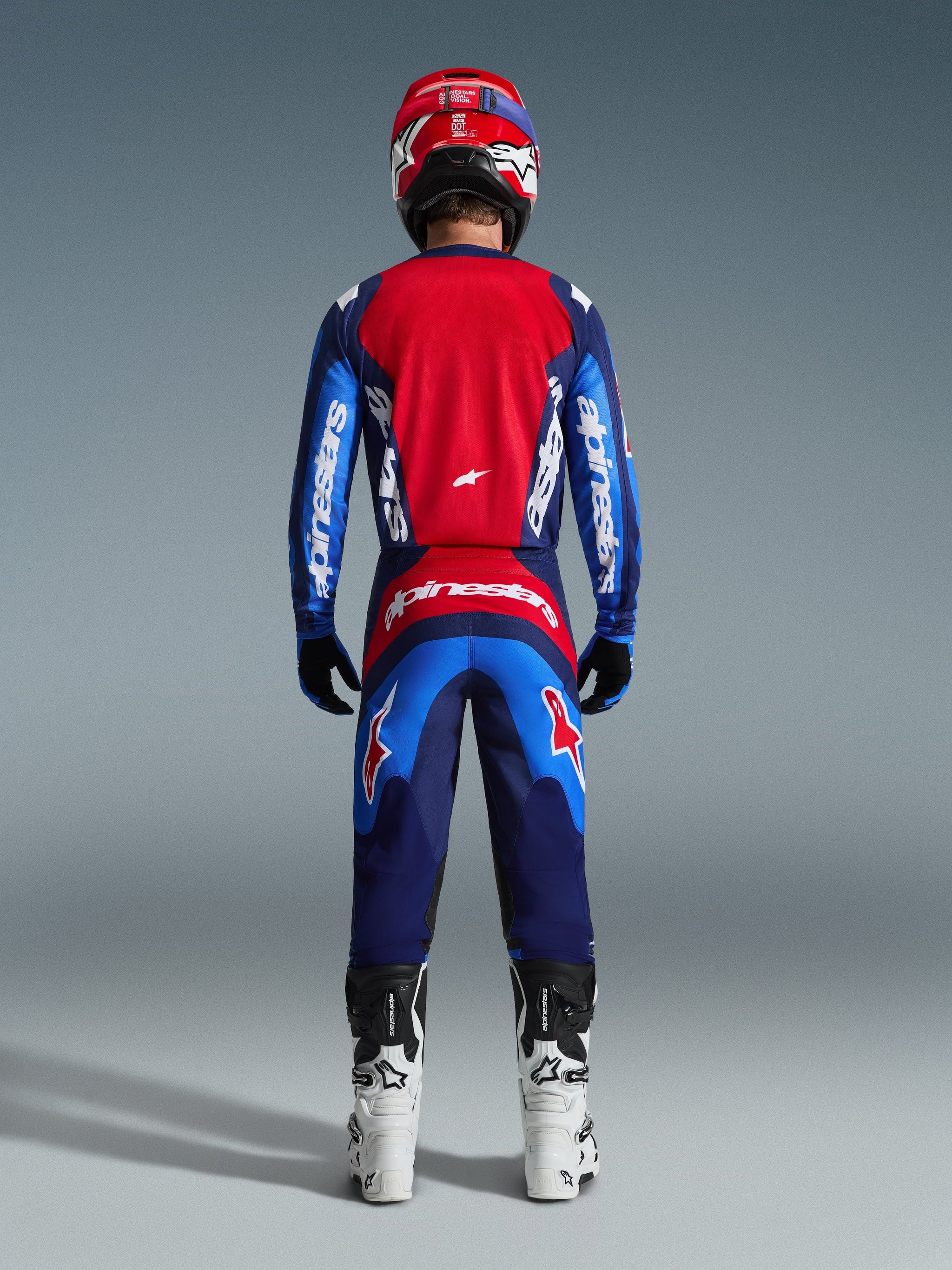 Motocross-Fahrer von hinten in kompletter Rennausrüstung, 2026 Fluid Grid Hose in Blau-Rot, passendes rot-blaues Trikot mit Alpinestars-Branding, rot-blauer Helm, weiß-schwarze Stiefel, abgestimmtes Outfit für Offroad-Rennsport