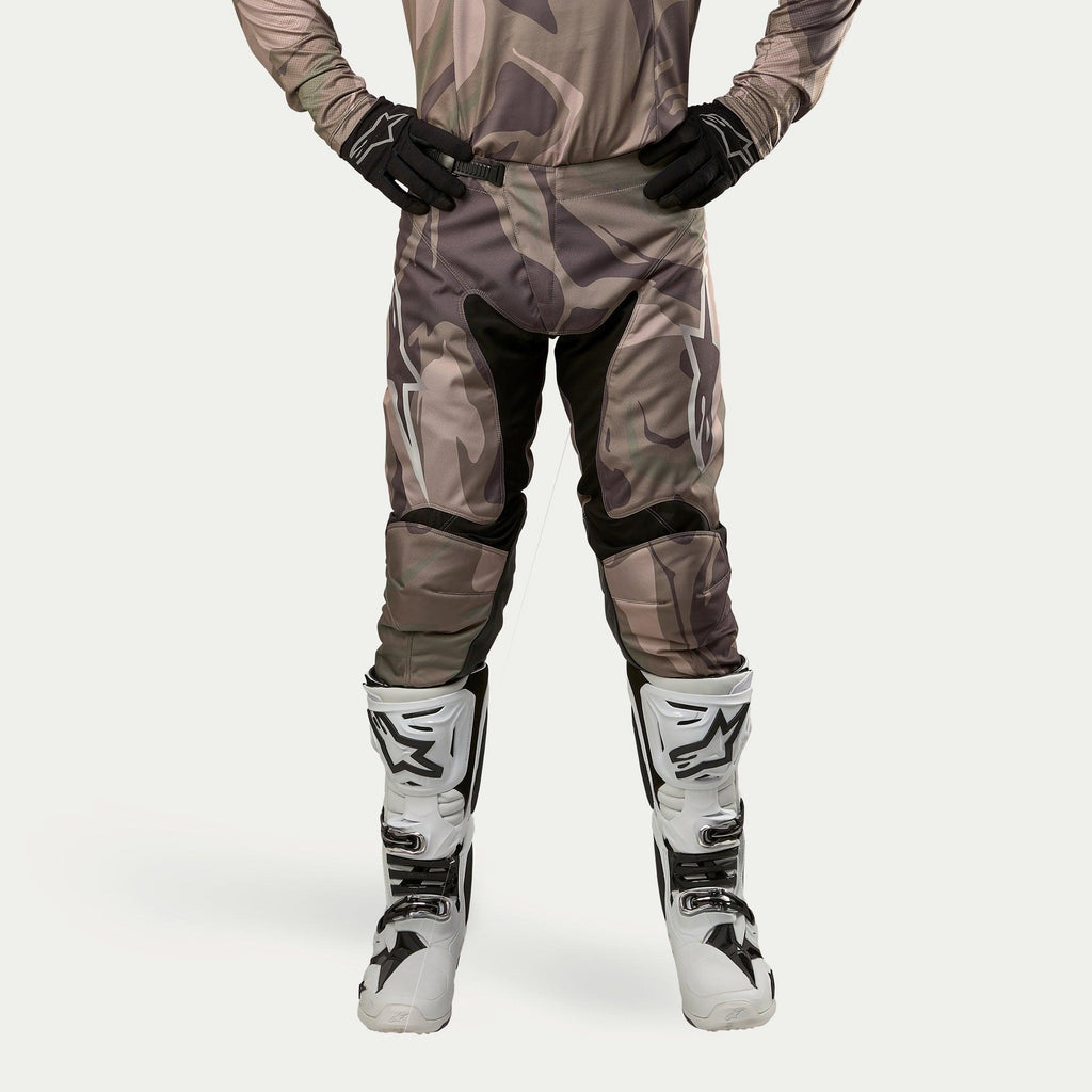 Alpinestars 2024 Racer Tactical Hose, Motocross-Hose, Military Green Camo Brown, mit großen weißen Alpinestars-Logos an den Oberschenkeln und schwarzen Innenbein-Einsätzen, getragen mit weiß-schwarzen Motocross-Stiefeln und schwarzen Handschuhen, passendes Camouflage-Trikot, Frontansicht.