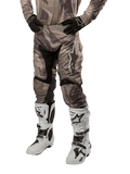 Alpinestars 2024 Racer Tactical Hose, Motocross-Hose, Militärgrün Camo Braun, getragen mit passendem Camouflage-Trikot, schwarzen Handschuhen und weißen Stiefeln mit Stern-Logos, mit schwarzen Stretch-Einsätzen und ergonomischem Design, Offroad-Ausrüstung auf weißem Hintergrund