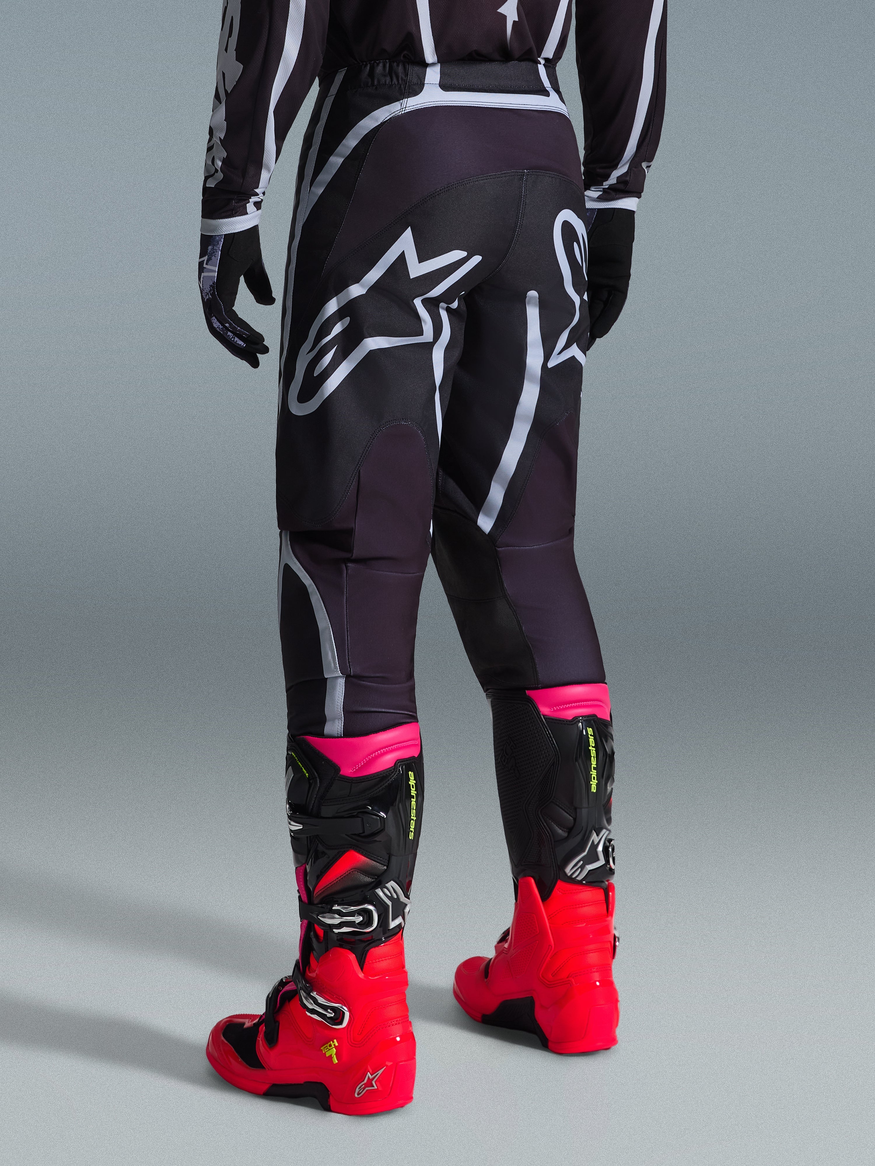 Alpinestars 2026 Fluid Apex Pants, Motocross Pants, Black and Gray, rear view featuring large white Alpinestars logos on the seat, worn with bright red and pink boots, durable 600D polyfabric construction for off-road performance