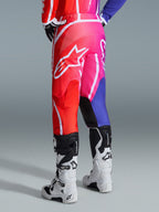 Alpinestars 2026 Fluid Apex Hose, Motocross-Hose, Mehrfarbig in Rot, Pink und Lila, Rückansicht eines Fahrers mit weiß-schwarzen Motocross-Stiefeln, großes weißes Alpinestars-Logo, lebendiges Farbverlaufs-Design