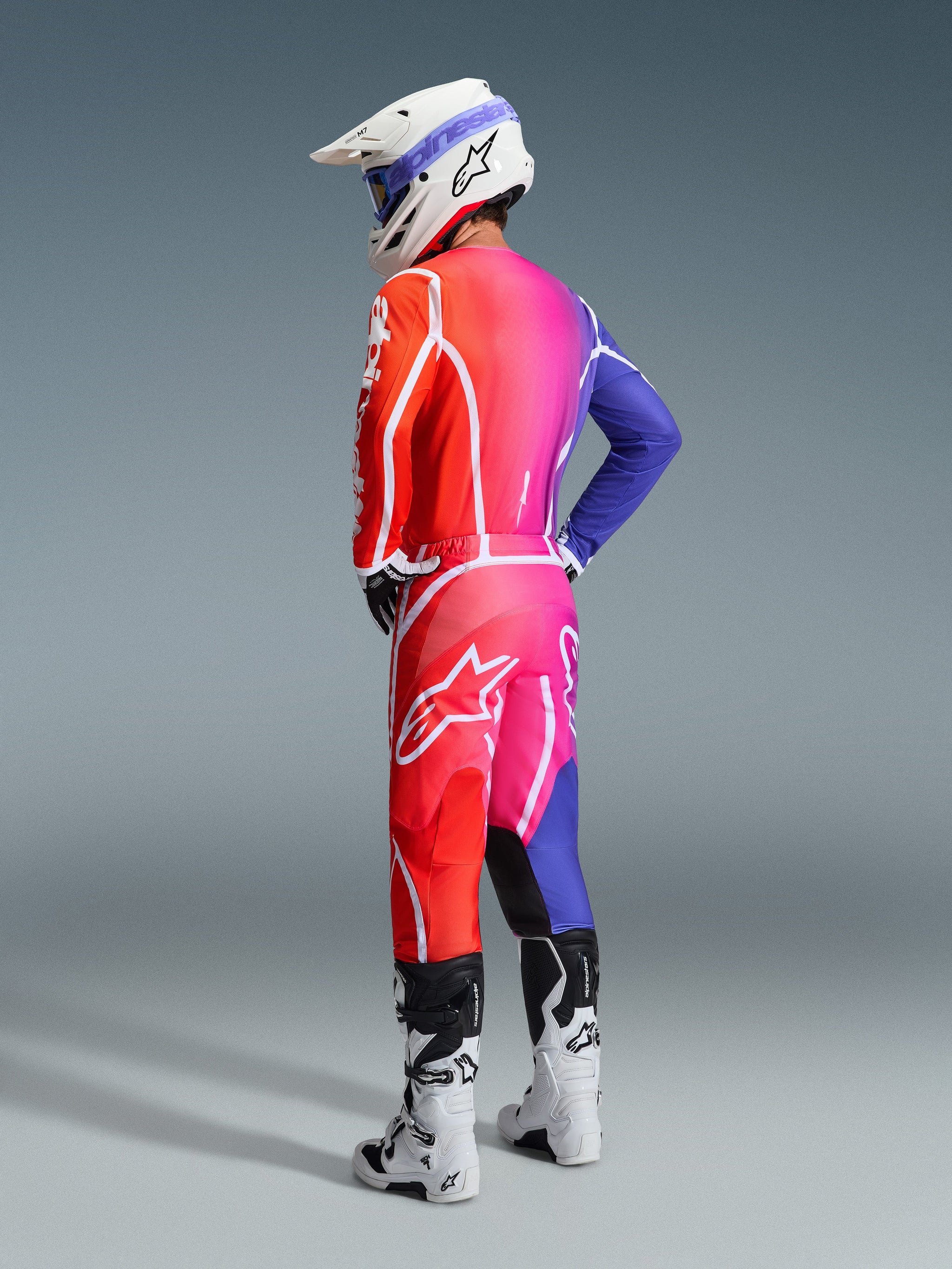 Motocross rider wearing Alpinestars 2026 Fluid Apex gear, Multicolor jersey and pants featuring a red, pink, and purple gradient with white logos, white helmet and white boots, coordinated outfit for off-road racing