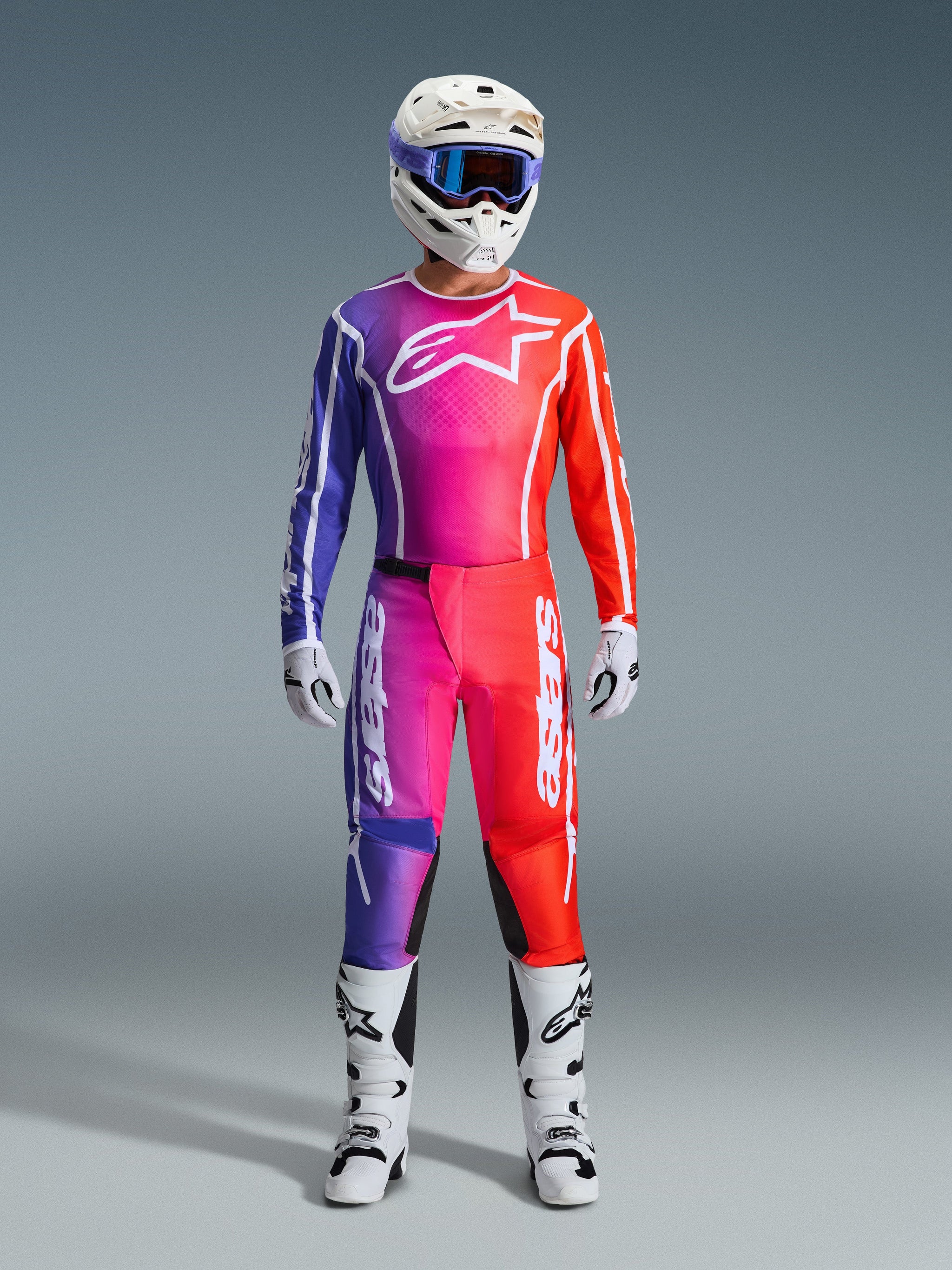 Motocross rider wearing 2026 Fluid Apex Pants and matching jersey, purple and orange gradient, white helmet with goggles, white boots and gloves, coordinated Alpinestars racing gear for off-road performance.