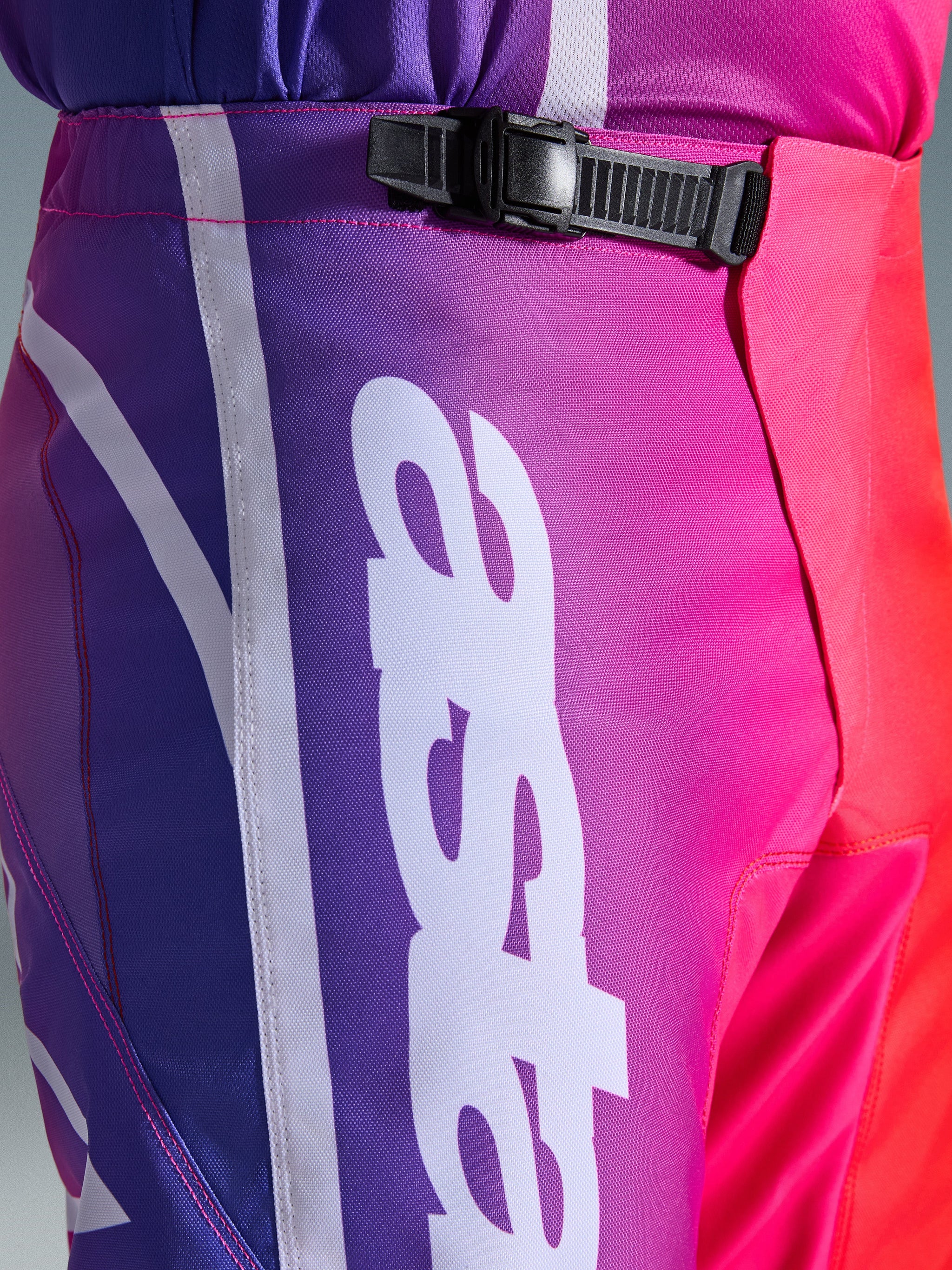 Alpinestars 2026 Fluid Apex Pants, Motocross Pants, Multicolor Purple Pink and Red, close-up of the waist and hip area featuring a black ratchet closure system and large white vertical Alpinestars logo, vibrant gradient design with durable polyfabric texture