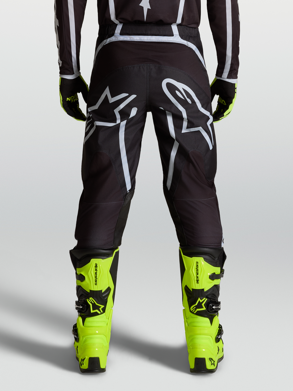 Alpinestars Fluid Apex V2 Hose, Motocross-Hose in Schwarz und Silber, Rückansicht mit ergonomischer Passform, verstärkten Bereichen und großem Astars-Logo, getragen mit neongelben Tech 7 Stiefeln für Offroad-Performance.