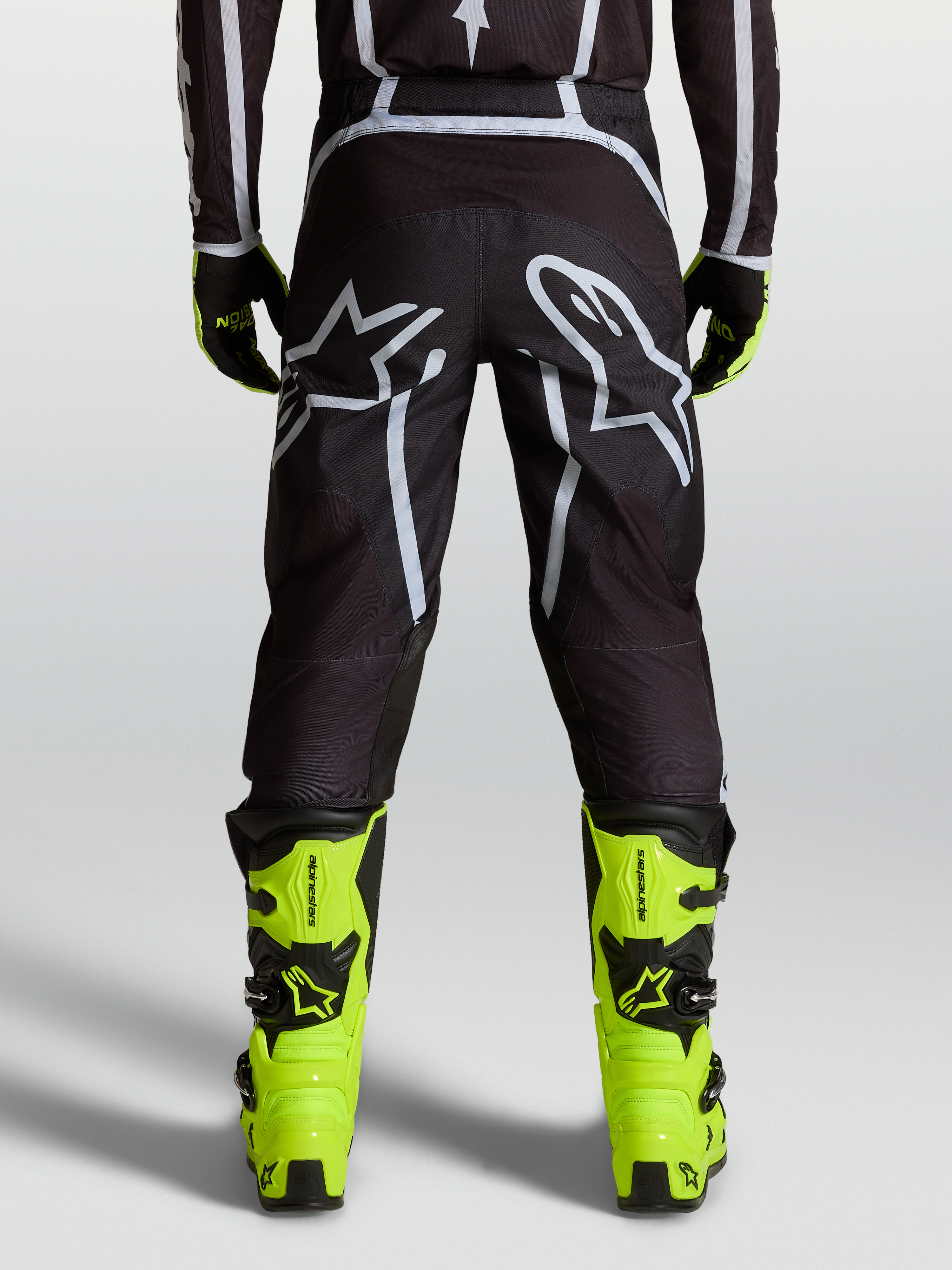 Alpinestars Fluid Apex V2 Hose, Motocross-Hose in Schwarz und Silber, Rückansicht mit ergonomischer Passform, verstärkten Bereichen und großem Astars-Logo, getragen mit neongelben Tech 7 Stiefeln für Offroad-Performance.