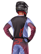 Motocross-Fahrer von hinten, trägt Alpinestars 2024 Racer Hoen Ausrüstung, Trikot in Lavan Purple und Schwarz mit rotem Logo, passende Hose mit roten Akzenten, schwarzer Helm, abgestimmtes Outfit für Offroad-Rennen