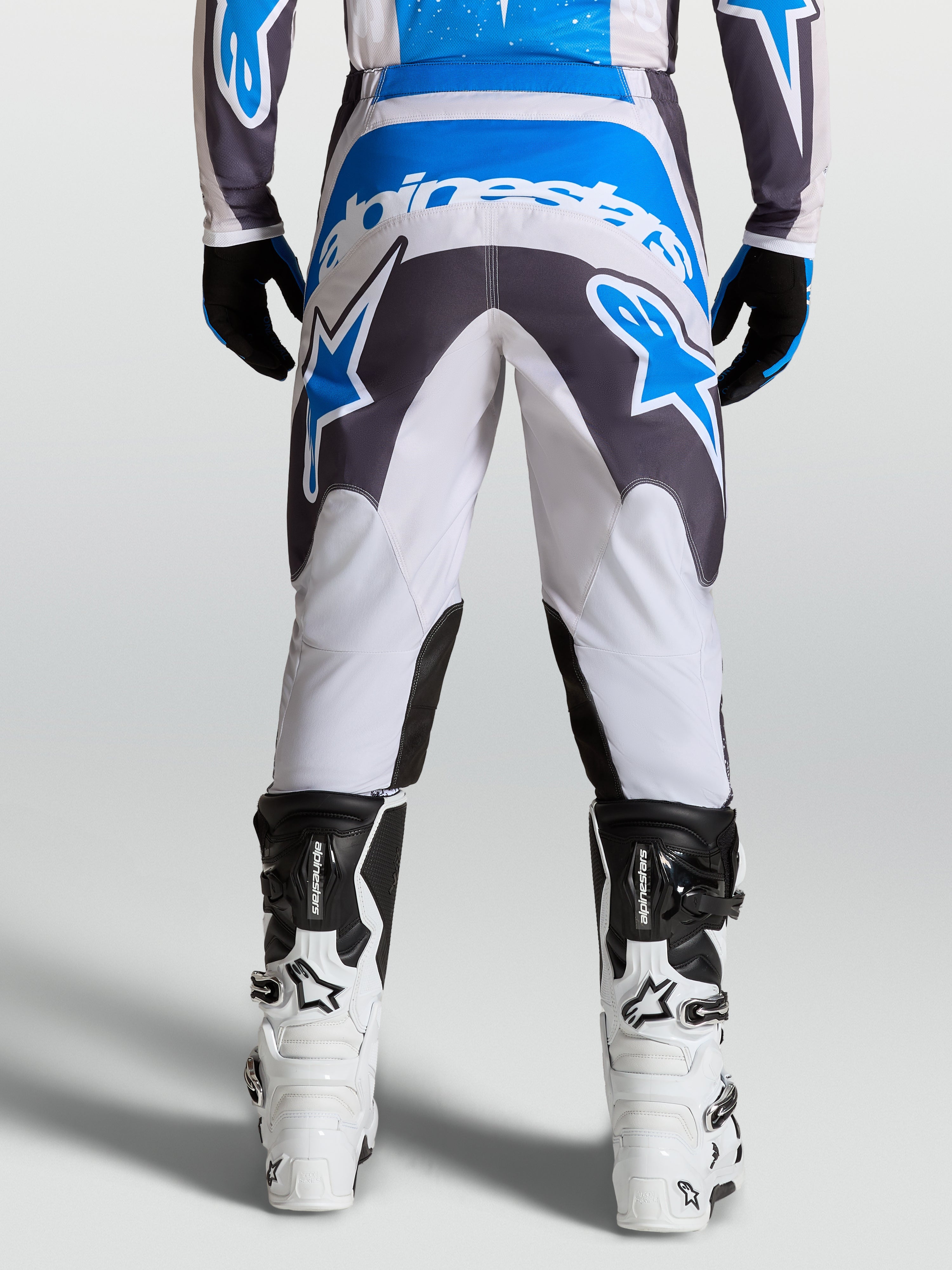 Alpinestars Fluid Drag Hosen, Herren-Motocross-Hose in Grau und Blau, verstärkter Sitzbereich für Offroad-Fahrten, Rückansicht am Fahrer mit weißen Motorradstiefeln.