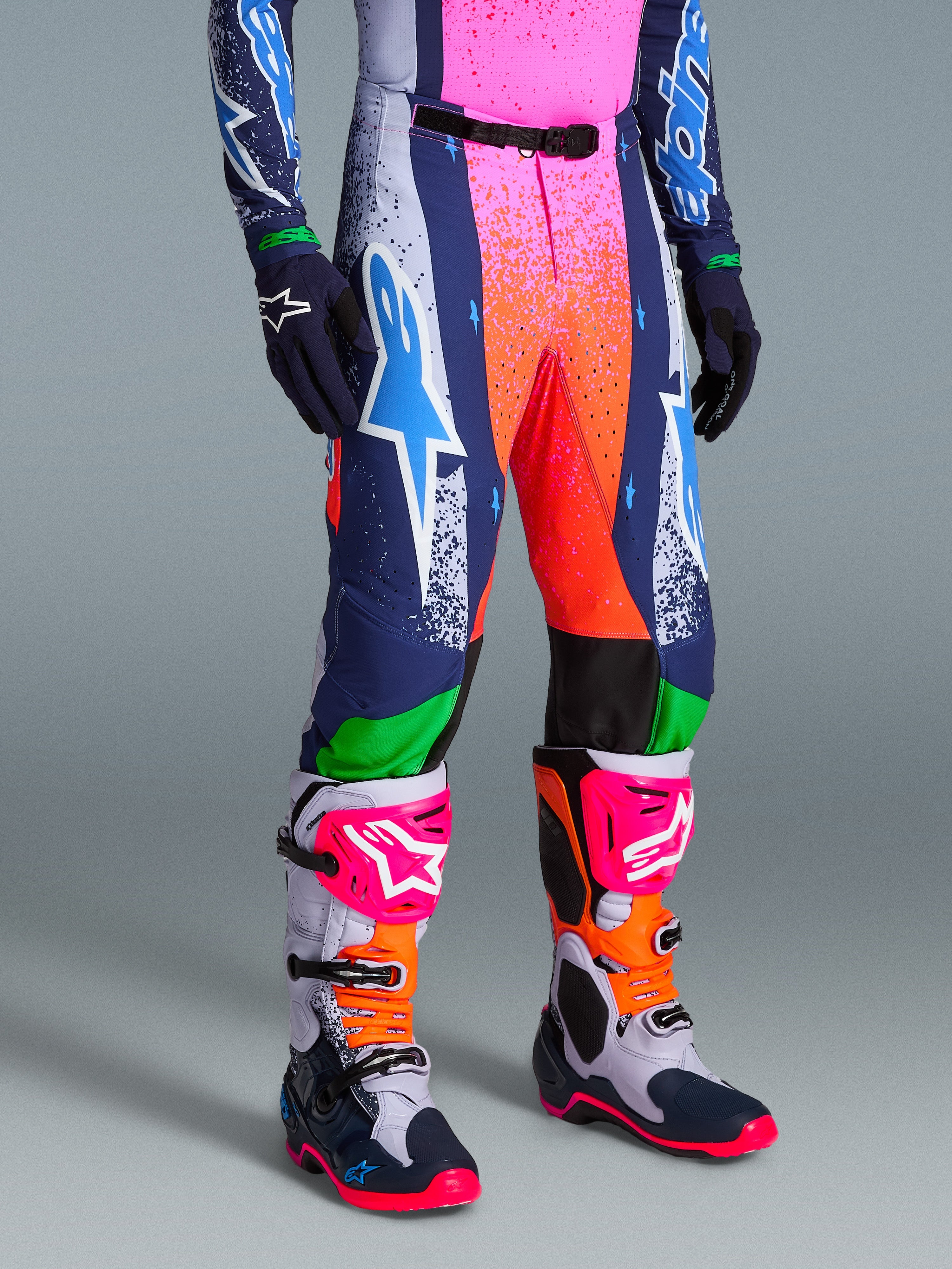 Alpinestars Limited Edition Supertech Records Hose, lebhafte mehrfarbige Motocross-Hose mit gesprenkelten Grafiken in Pink und Orange, dunkelblauen und weißen Einsätzen, mit einem großen weißen und cyanfarbenen Astars-Logo, getragen mit farblich abgestimmten Performance-Stiefeln und Handschuhen.