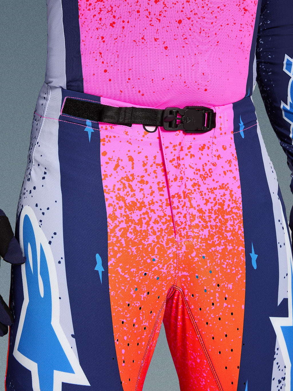 Alpinestars Limited Edition Supertech Records Hose, Motocross-Hose, Mehrfarbig Pink und Orange mit marineblauen Seiteneinsätzen, Nahaufnahme des Hosenbundes mit schwarzer, selbstverriegelnder SX1-Magnetschnalle, Ripstop-Gewebe mit Splatter-Grafiken und lasergeschnittenen Perforationen.