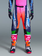 Alpinestars Limited Edition Supertech Records Hose, Motocross-Hose, Mehrfarbig mit lebendigen pink- und orangefarbenen Splash-Grafiken, dunkelblaue Einsätze mit blauen Stern-Logos und grünen Akzenten, kombiniert mit passendem Jersey sowie grau, pink und orangefarbenen Motocross-Stiefeln.