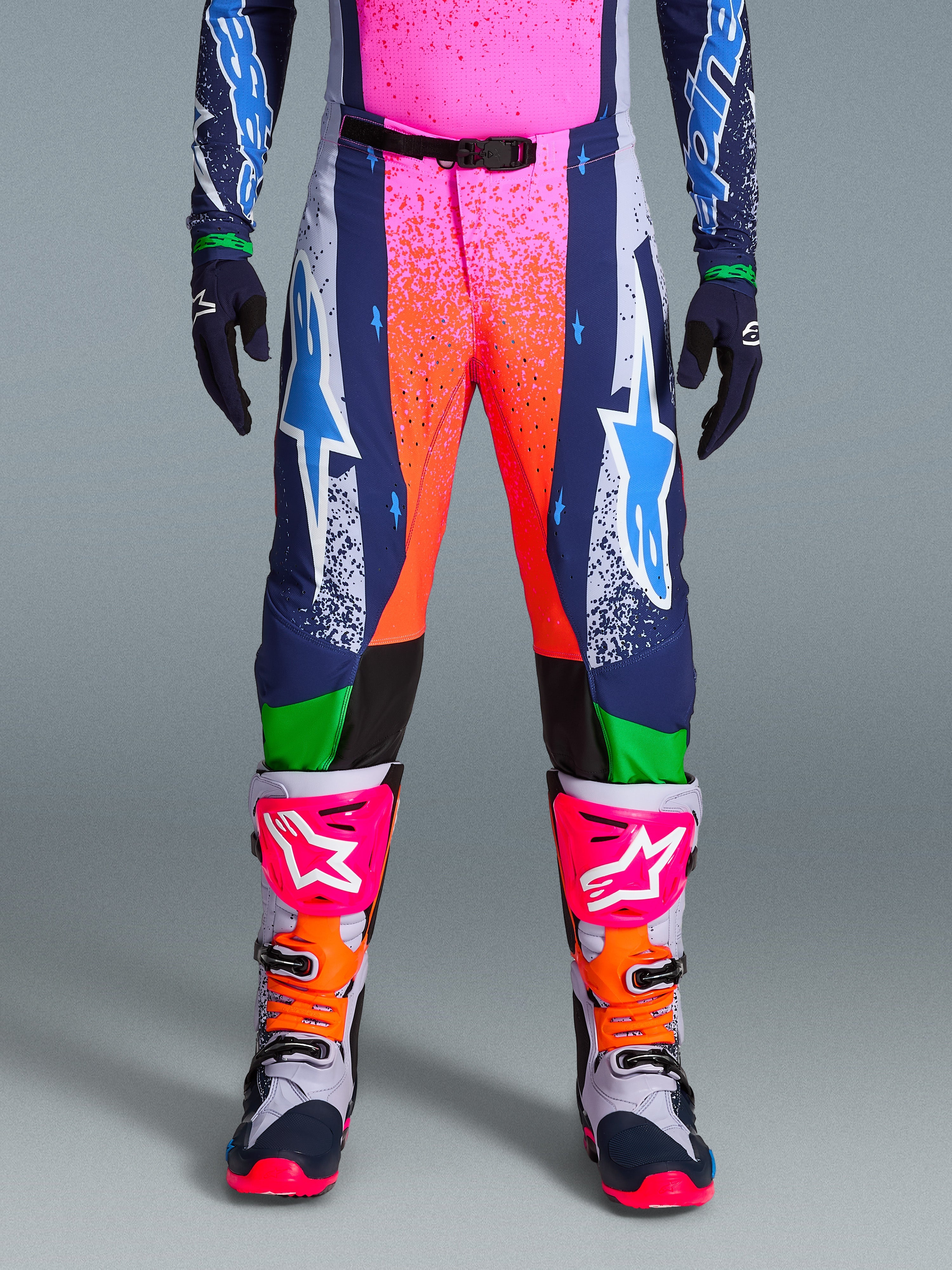 Alpinestars Limited Edition Supertech Records Hose, Motocross-Hose, Mehrfarbig mit lebendigen pink- und orangefarbenen Splash-Grafiken, dunkelblaue Einsätze mit blauen Stern-Logos und grünen Akzenten, kombiniert mit passendem Jersey sowie grau, pink und orangefarbenen Motocross-Stiefeln.