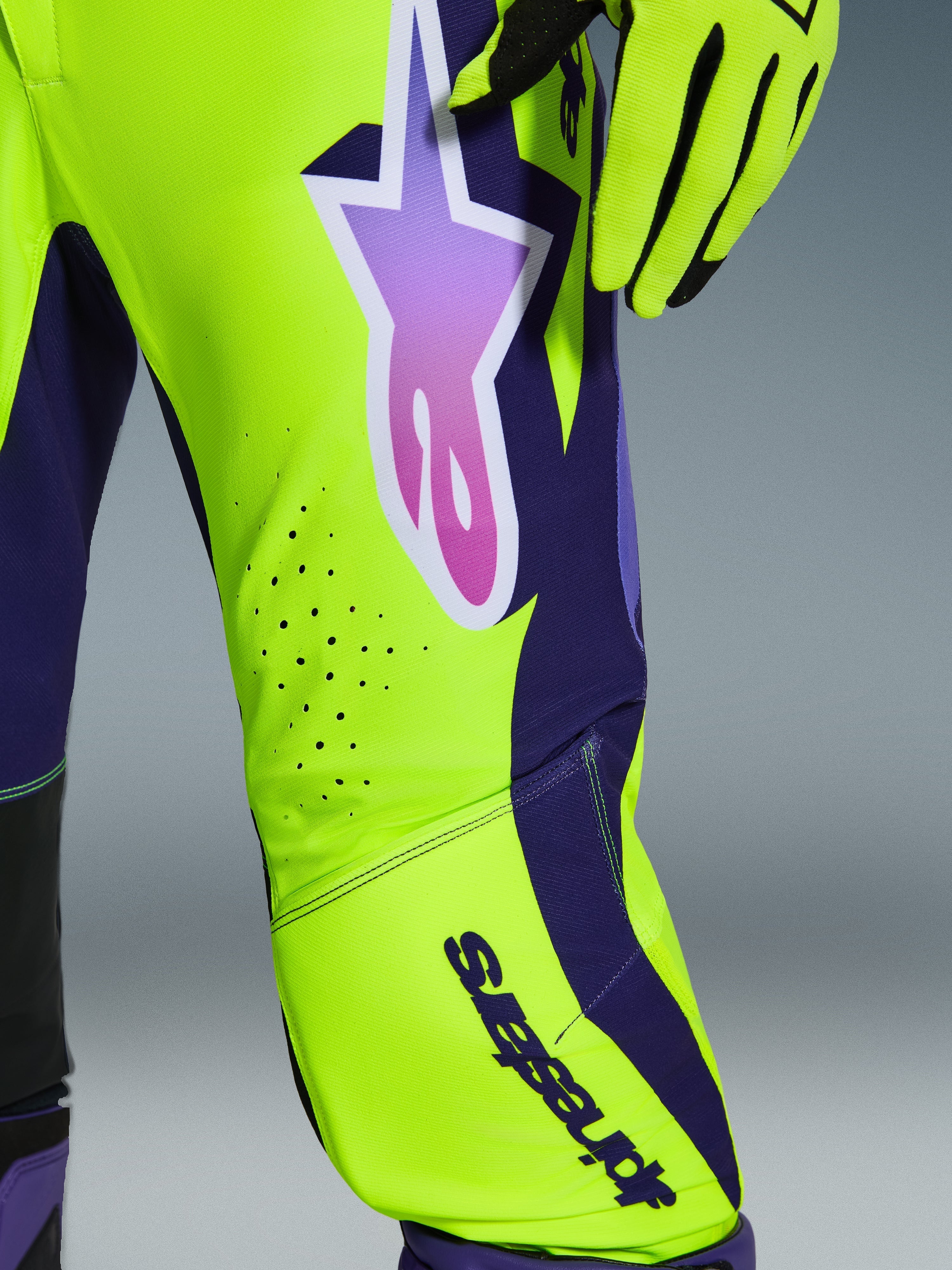 Alpinestars 2026 Supertech Vista Hose, Motocross-Hose, Gelb, Lila und Schwarz, Nahaufnahme mit Stern-Logo im Farbverlauf und lasergeschnittenen Belüftungslöchern, kombiniert mit passenden Rennhandschuhen