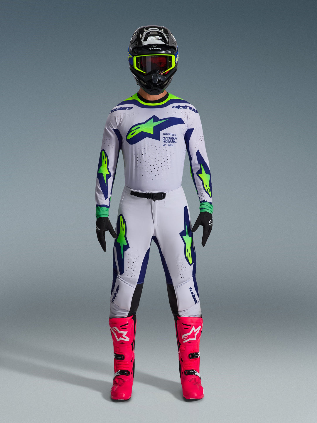 Alpinestars 2026 Supertech Vista Gear Set, Motocross-Fahrer in kompletter Rennausrüstung, weißes Jersey und Hose mit blauen und neongrünen Akzenten, schwarzer Helm mit neongelben Details, leuchtend pinke Stiefel, professionelles Offroad-Racing-Ensemble