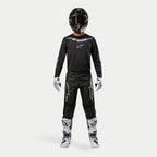Motocross rider wearing complete racing gear, Alpinestars 2024 Fluid Graphite Pants in Black and Silver, black long-sleeve jersey with silver branding, black helmet with goggles, white and black boots, white and black gloves, coordinated outfit for motocross