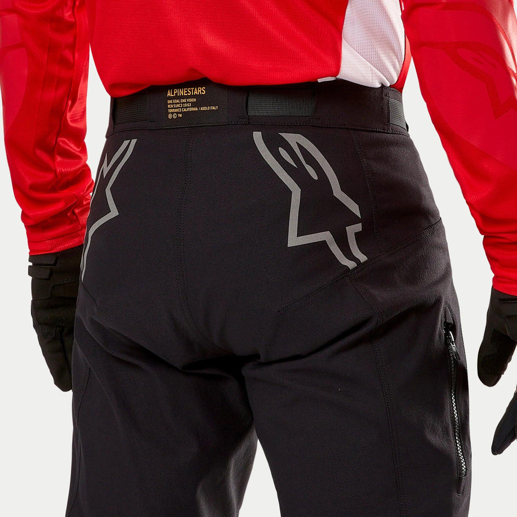 Alpinestars Techdura Hose, schwarze Enduro-Hose, Rückansicht mit großen grauen Astars-Logos auf dem Gesäß, getragen mit einem roten Trikot und schwarzen Handschuhen, strapazierfähiges Material mit seitlichem Reißverschluss und Marken-Bund