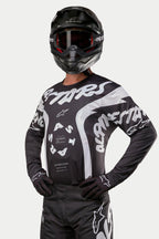 Alpinestars 2024 Racer Hana Jersey, Motocross-Jersey, Schwarz und Weiß, mit auffälligen weißen Alpinestars-Textgrafiken und Logo, getragen von einem Fahrer mit schwarzem Helm und Brille, Hochleistungs-Ausrüstung für den Offroad-Rennsport