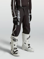 Alpinestars Jugend Fluid Apex V2 Hose, Motocross-Hose für junge Fahrer in Schwarz und Silber, ergonomisch vorgeformte Passform, getragen mit weißen Tech-Stiefeln.