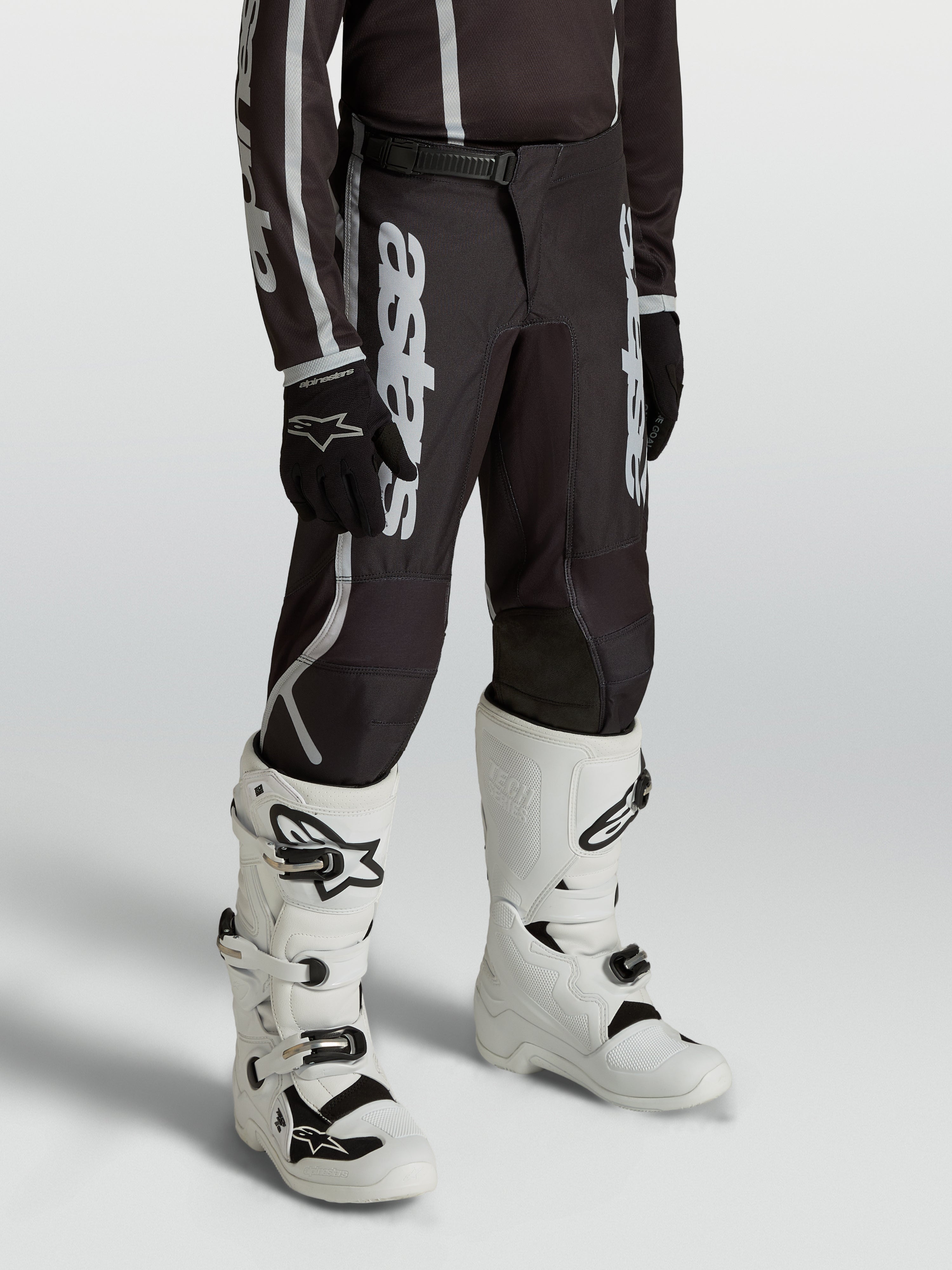 Alpinestars Jugend Fluid Apex V2 Hose, Motocross-Hose für junge Fahrer in Schwarz und Silber, ergonomisch vorgeformte Passform, getragen mit weißen Tech-Stiefeln.