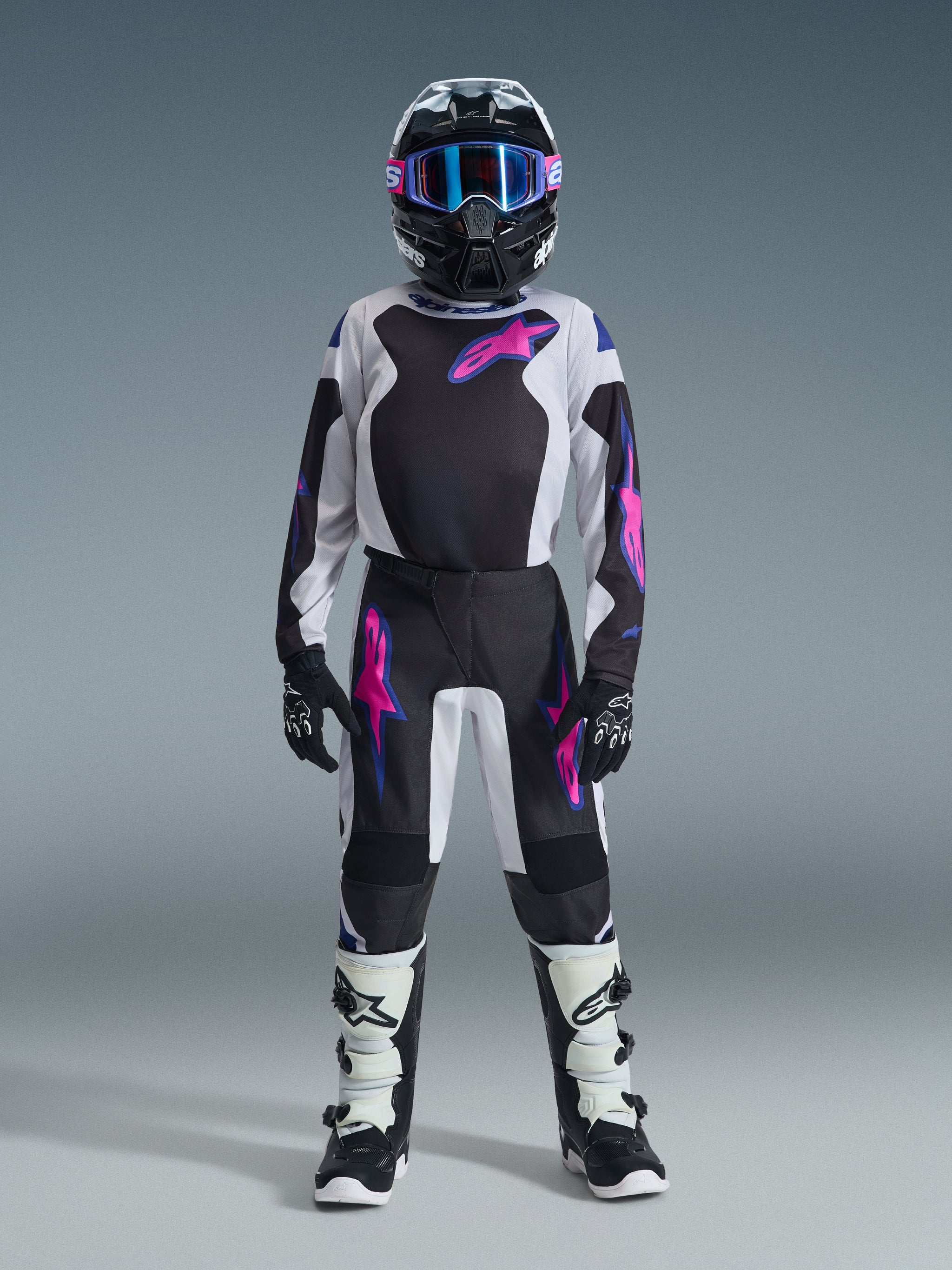 Jugendlicher Motocross-Fahrer in Alpinestars 2026 Youth Fluid Grid Hose in Schwarz, Hellgrau und Lila, passendes langärmeliges Trikot, schwarzer Helm mit pink-lila Brille, weiß-schwarze Stiefel und schwarze Handschuhe, komplett abgestimmtes Ausrüstungsset für den Offroad-Einsatz.