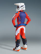 Youth motocross rider viewed from behind wearing Alpinestars 2026 Youth Fluid Grid Pants in Orange and Ucla Blue, matching orange and blue jersey, white helmet with goggles, and white and orange motocross boots, complete coordinated outfit for off-road racing.