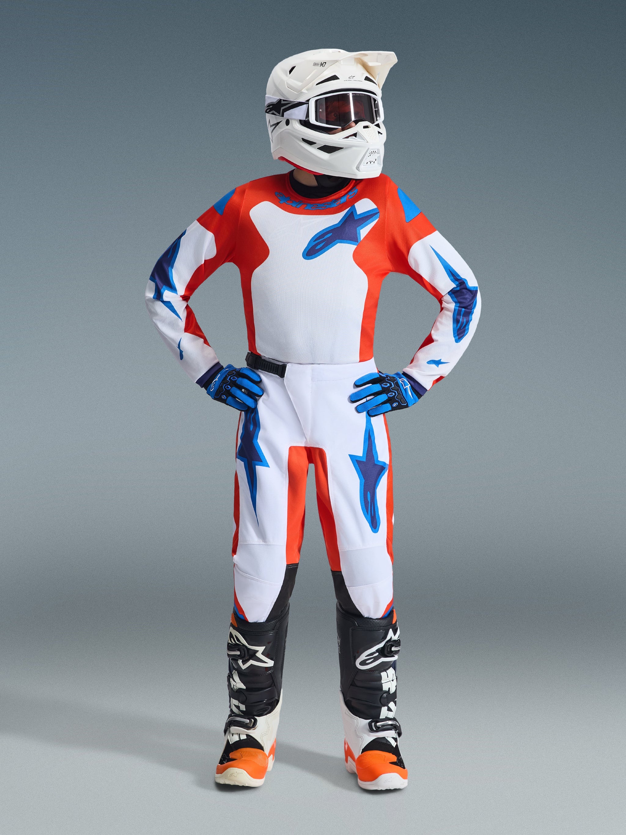 Youth motocross rider wearing complete racing gear, Alpinestars 2026 Youth Fluid Grid Pants in Orange and UCLA Blue, matching white and orange jersey with blue star logos, white helmet, blue gloves, black and white boots, coordinated outfit for off-road racing