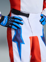 Alpinestars 2026 Youth Fluid Grid Pants, Motocross Pants, Orange and UCLA Blue, white base with orange side panels and blue star graphics, featuring a black ratchet waist closure, worn with matching blue and black gloves, professional gear for youth off-road racing.