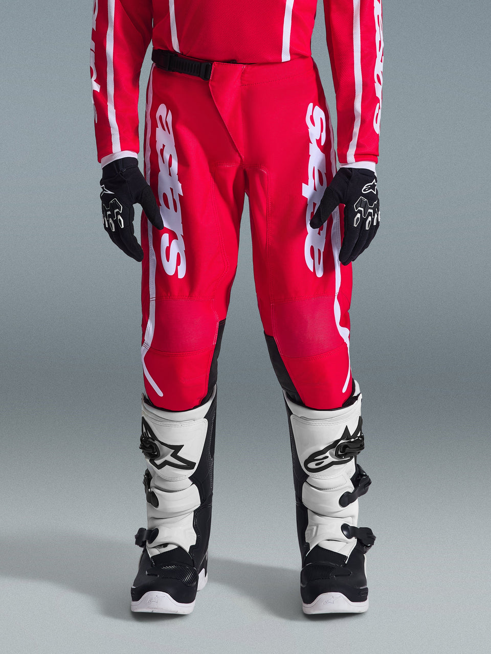 Alpinestars 2026 Youth Fluid Apex Pants, Motocross Pants, Red and White, shown on a rider with matching red jersey, black gloves, and white and black boots, featuring large white vertical Alpinestars branding and black inner knee reinforcement for off-road performance