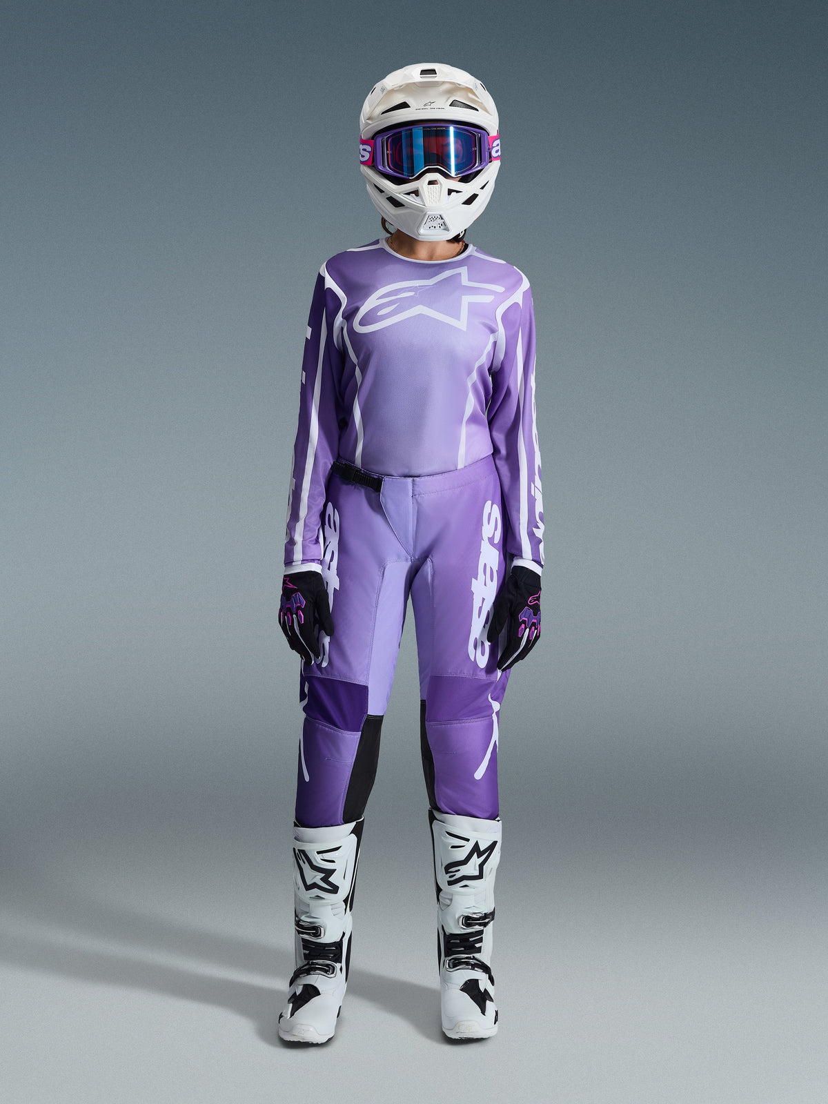Motocross-Fahrerin im Alpinestars 2026 Stella Fluid Apex Bekleidungsset für Damen, violett-weißes Jersey und passende Hose, weißer Helm mit pinker Brille, weiß-schwarze Offroad-Stiefel, vollständige Rennausrüstung für Damen-Motocross.