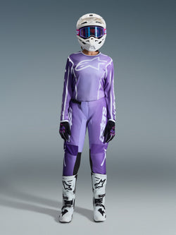 Motocross rider wearing the Alpinestars 2026 Woman Stella Fluid Apex Gear Set, violet and white jersey with matching pants, white helmet with pink goggles, and white and black off-road boots, complete racing kit for women's motocross.