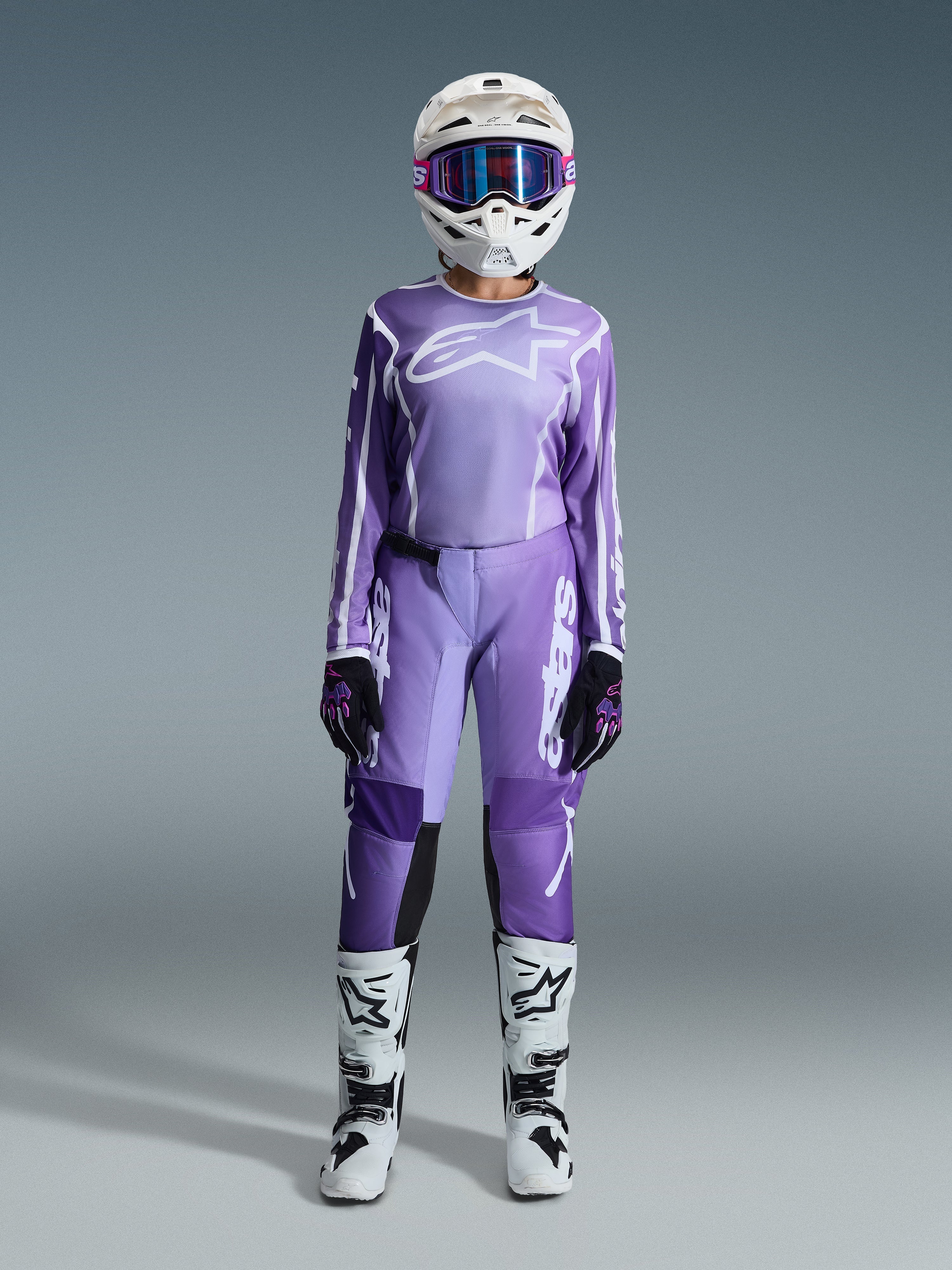 Motocross-Fahrerin im Alpinestars 2026 Stella Fluid Apex Bekleidungsset für Damen, violett-weißes Jersey und passende Hose, weißer Helm mit pinker Brille, weiß-schwarze Offroad-Stiefel, vollständige Rennausrüstung für Damen-Motocross.
