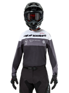 Motocross-Fahrer im Alpinestars Pro-Dura Jersey in Black Magnet White, langärmliges Performance-Oberteil mit weißen und grauen Farbblöcken, kombiniert mit schwarzem Helm und Schutzbrille, Alpinestars-Branding und „ONE GOAL ONE VISION“-Slogan, Offroad-Motorrad-Rennbekleidung