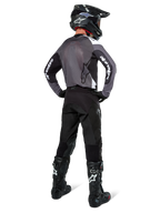 Motocross-Fahrer im Alpinestars Pro-Dura Trikot in Black Magnet White, Rückansicht mit grauem Mesh-Rückenteil und schwarzen Ärmeln mit weißem Alpinestars-Branding, kombiniert mit schwarzer Hose, schwarzem Helm und schwarz-weißen Stiefeln, professionelle Enduro-Rennausrüstung
