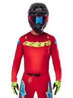 Alpinestars 2025 Supertech Maker Jersey in leuchtendem Rot und Gelb Fluo, mit gelbem „astars“-Branding auf der Brust und blauen Stern-Designs an den Schultern, getragen mit passender Hose, schwarzem Helm, blauer Brille und gelben Handschuhen, professionelle Motocross-Rennbekleidung
