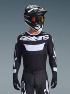 Alpinestars 2026 Racer Air Riway Jersey, schwarz-weißes Motocross-Trikot mit 'astars'-Branding, getragen von einem Fahrer mit passendem schwarzem Helm, Goggles und Hose, mit atmungsaktiven Mesh-Einsätzen für den Offroad-Rennsport.