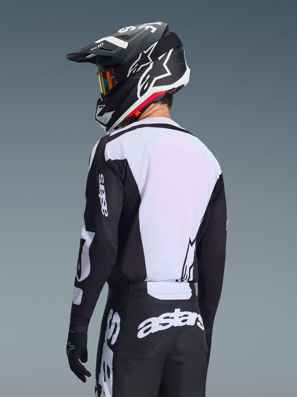 Motocross-Fahrer von hinten im Alpinestars 2026 Racer Air Riway Jersey in Schwarz und Weiß, mit weißem, atmungsaktivem Mesh-Rückenpanel und schwarzen Ärmeln mit „astars“-Branding, passender schwarzer Hose mit weißen Logos und schwarzem Helm mit weißen Akzenten, professionelle Offroad-Ausrüstung.