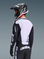 Motocross-Fahrer von hinten im Alpinestars 2026 Racer Air Riway Jersey in Schwarz und Weiß, mit weißem, atmungsaktivem Mesh-Rückenpanel und schwarzen Ärmeln mit „astars“-Branding, passender schwarzer Hose mit weißen Logos und schwarzem Helm mit weißen Akzenten, professionelle Offroad-Ausrüstung.