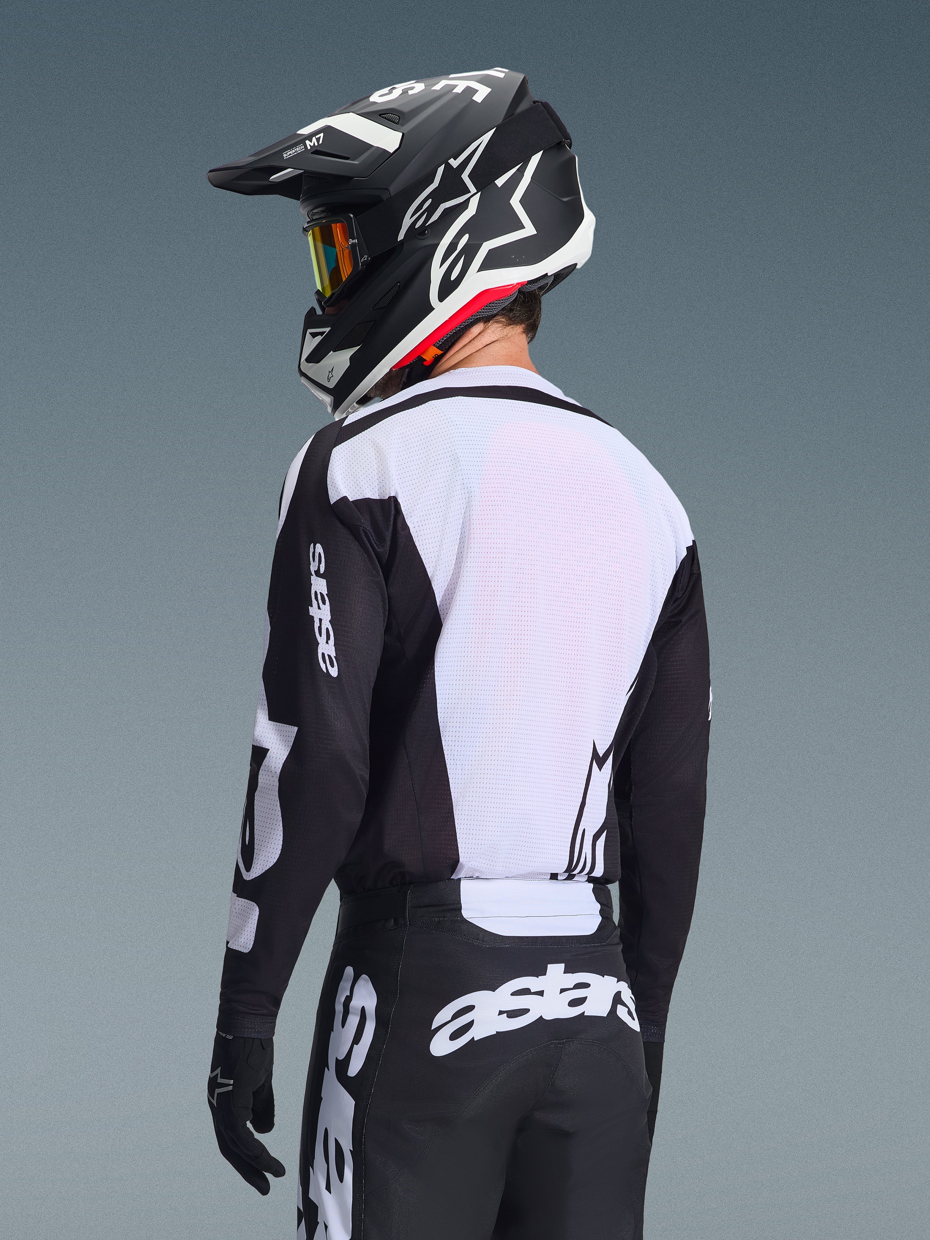 Motocross-Fahrer von hinten im Alpinestars 2026 Racer Air Riway Jersey in Schwarz und Weiß, mit weißem, atmungsaktivem Mesh-Rückenpanel und schwarzen Ärmeln mit „astars“-Branding, passender schwarzer Hose mit weißen Logos und schwarzem Helm mit weißen Akzenten, professionelle Offroad-Ausrüstung.