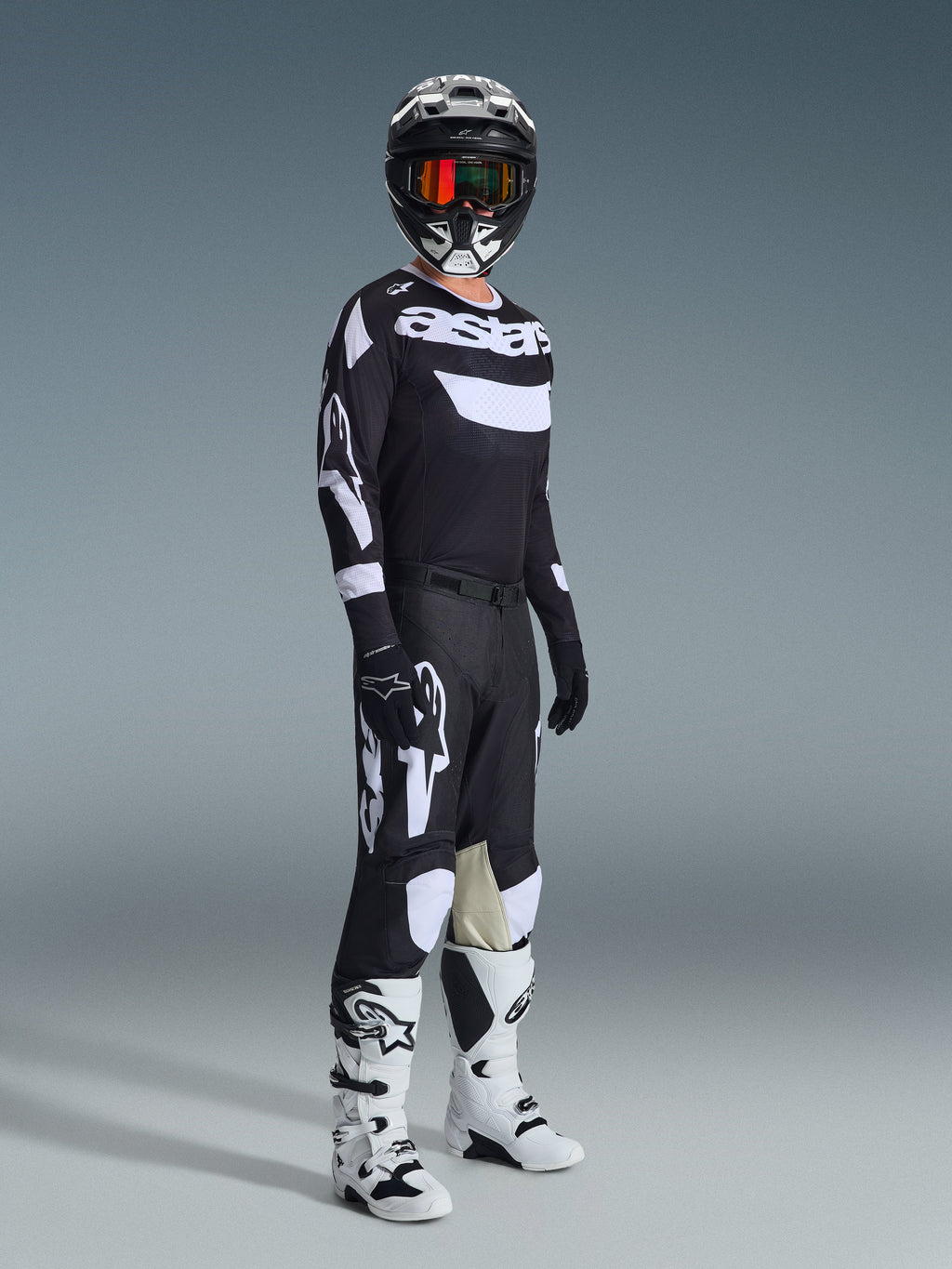 Motocross-Fahrer in kompletter Rennausrüstung, 2026 Racer Air Riway Trikot in Schwarz und Weiß, passende schwarze Hose mit großen weißen Alpinestars-Logos, schwarzer Helm mit weißen Akzenten und orangefarbener Brille, weiß-schwarze Stiefel, abgestimmtes Outfit für professionellen Offroad-Rennsport