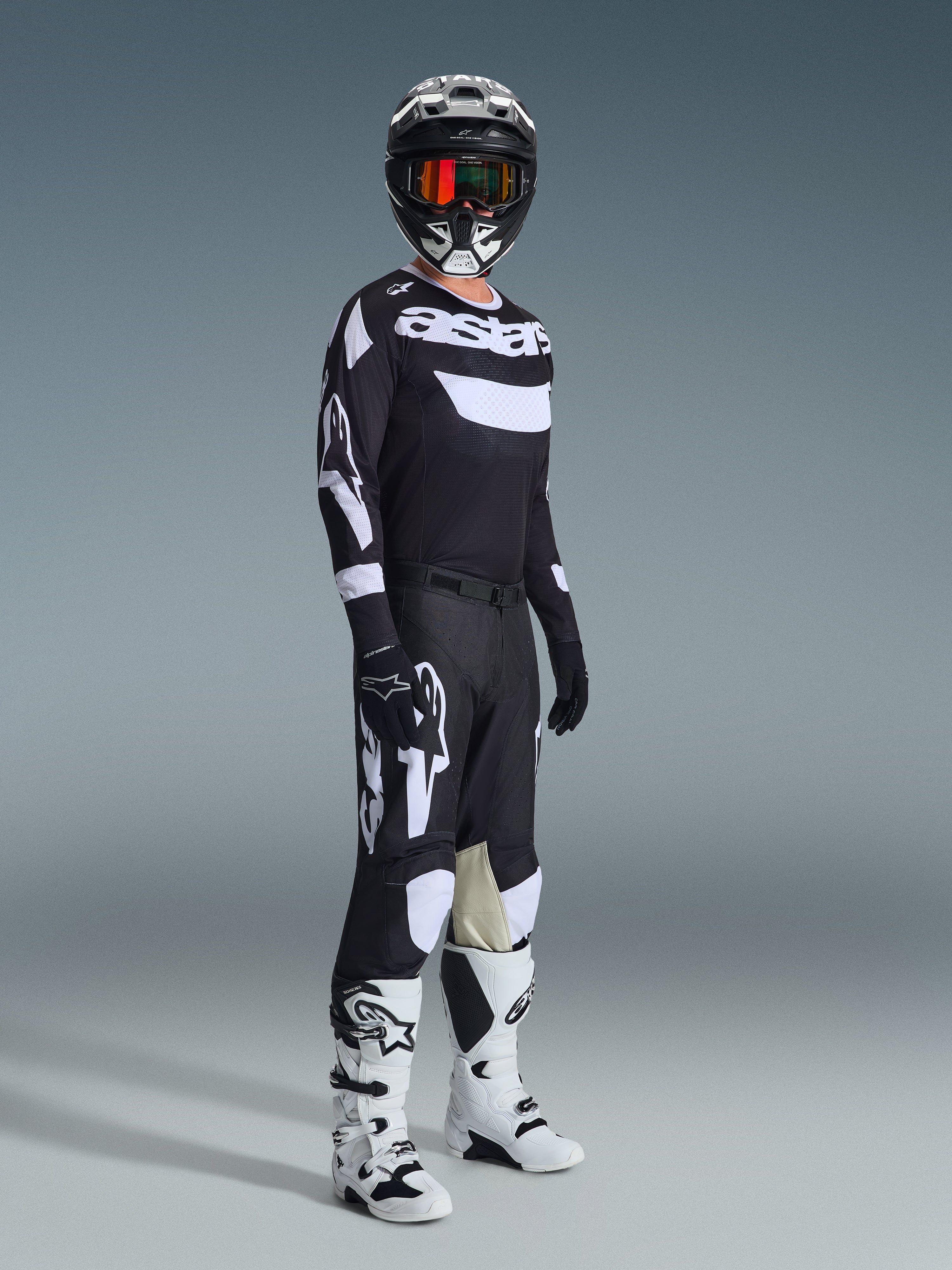 Motocross-Fahrer in kompletter Rennausrüstung, 2026 Racer Air Riway Trikot in Schwarz und Weiß, passende schwarze Hose mit großen weißen Alpinestars-Logos, schwarzer Helm mit weißen Akzenten und orangefarbener Brille, weiß-schwarze Stiefel, abgestimmtes Outfit für professionellen Offroad-Rennsport