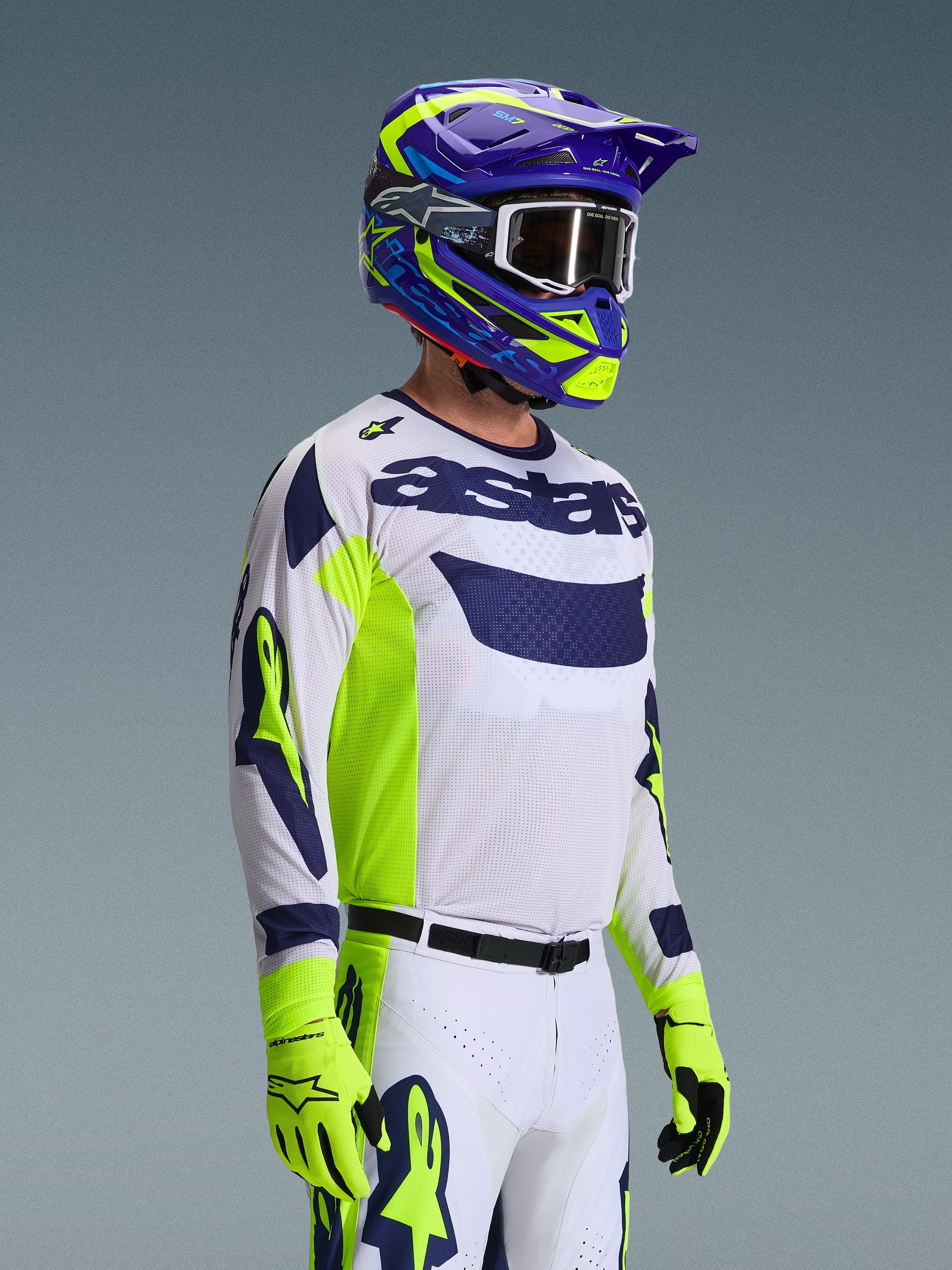 Motocross rider wearing the Alpinestars 2026 Racer Air Riway Jersey, Light Gray with Yellow Fluo and Blue accents, paired with a matching blue and yellow helmet and fluo yellow gloves, featuring breathable mesh construction and professional race styling for off-road performance.