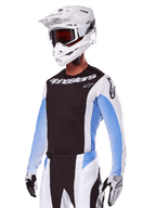 Motocross-Fahrer im Alpinestars 2025 Techstar Melt Gear Set, Weiß mit schwarzen und hellblauen Akzenten, Trikot mit schwarzer Brust und weißen Ärmeln, passender weiß-schwarzer Hose, weißem SM10 Helm mit Schutzbrille und schwarz-weißen Handschuhen, komplette professionelle Offroad-Rennausrüstung