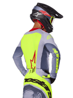 Alpinestars 2025 Techstar Melt Trikot, Bright Red Gray, Rückansicht eines Motocross-Fahrers in grauer und neongelber Rennausrüstung, rotes Stern-Logo auf dem Rücken, passender schwarz-roter Helm, Performance-Offroad-Bekleidung