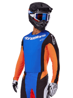 Alpinestars 2025 Techstar Melt Gear Set, Motocross-Fahrer in blauem Trikot mit orangefarbenen Ärmeln und schwarzen Seiteneinsätzen, passender blau-schwarzer Hose, schwarzer Helm mit blauer Brille, weiß-schwarze Handschuhe, sportliche Motocross-Bekleidung in Race-Passform
