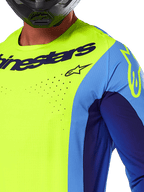 Alpinestars 2025 Techstar Melt Trikot, Motocross-Trikot, Gelb Fluo und Blau, mit lasergeschnittenen Perforationen und auffälligem Logo auf der Brust, athletische Passform für Offroad-Rennsport, Nahaufnahme mit schwarzem Helm