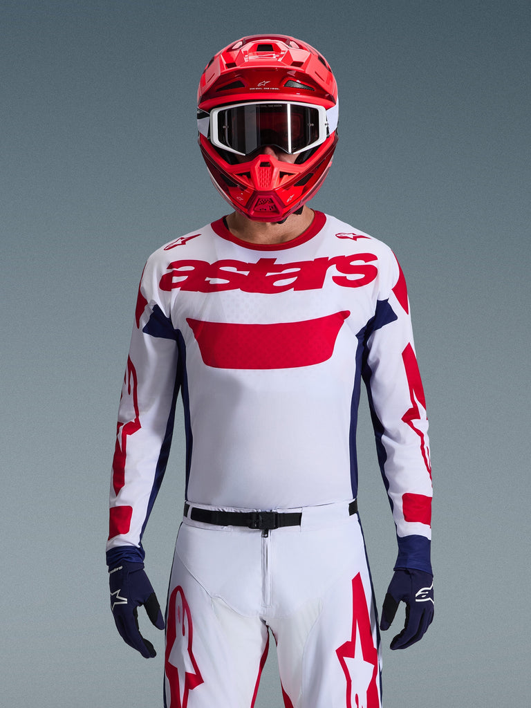 Motocross rider wearing the Alpinestars 2026 Racer Riway Jersey in White Blue Red, featuring a large red 'astars' logo on the chest, navy blue side panels, matching white and red motocross pants, a red helmet with white goggles, and navy blue gloves, complete off-road racing gear set.