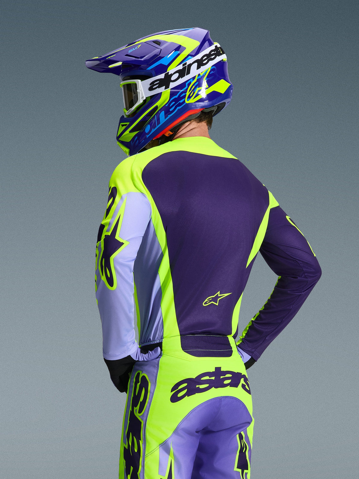 Motocross-Fahrer von hinten im Alpinestars 2026 Racer Portl Trikot und passender Hose, Farbschema in Lila und Gelb Fluo mit Lavendel-Akzenten, abgestimmter blau-gelber Helm, Rennpassform-Ausrüstung für Offroad-Performance