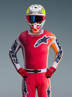 Alpinestars 2026 Racer Portl Trikot, rotes Motocross-Trikot, Vorderansicht eines Fahrers in passender roter Hose und Handschuhen, mit einem schwarz-weißen Stern-Logo auf der Brust und weißen Ärmeln mit schwarzem Branding, kombiniert mit einem mehrfarbigen weiß-gelben Helm, komplette professionelle Offroad-Rennausrüstung.